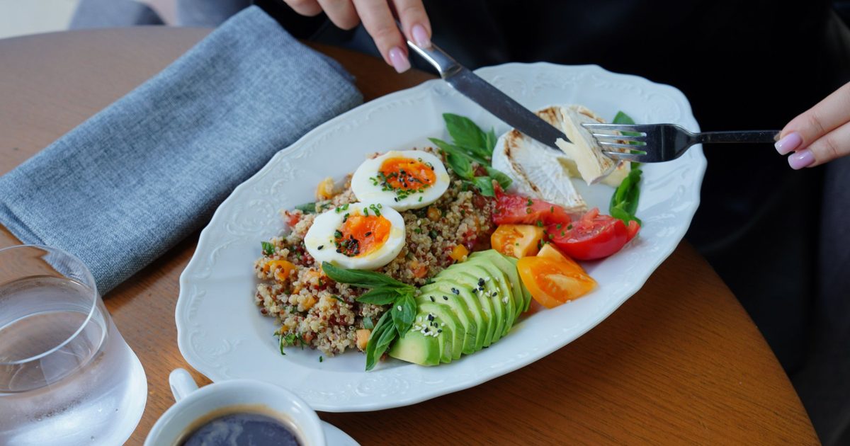 Persona cortando un plato con quinoa, huevo cocido, aguacate, tomate y queso, con agua y café en la mesa.