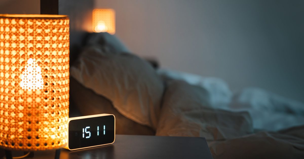Lámpara encendida junto a una cama, en un dormitorio en penumbra por la noche.
